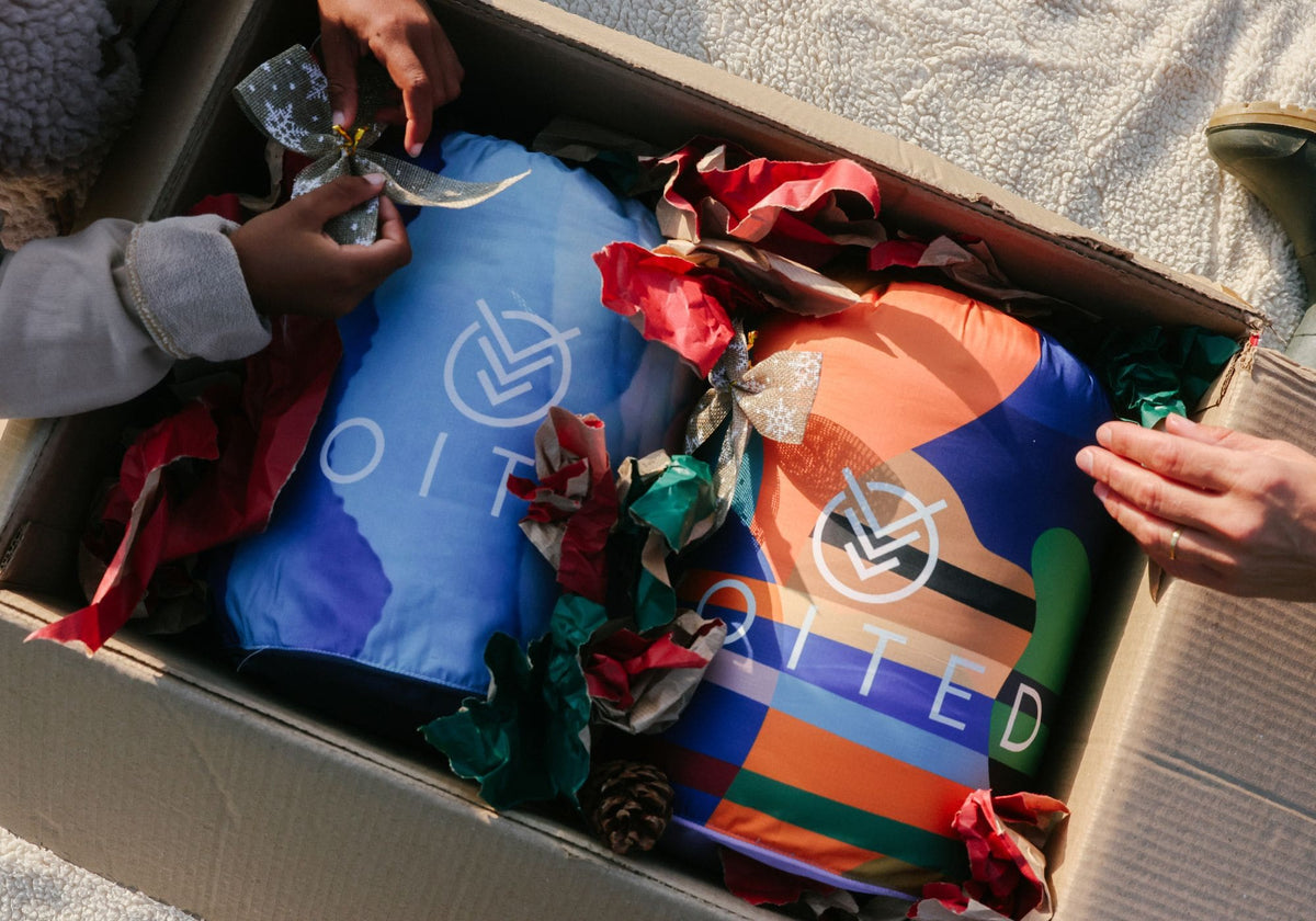 An open cardboard box containing two colorful insulated bags VOITED Cloud touch blankets , surrounded by red and green gift ribbon. Human hands are visible opening the box, indicating a lifestyle or unboxing context.