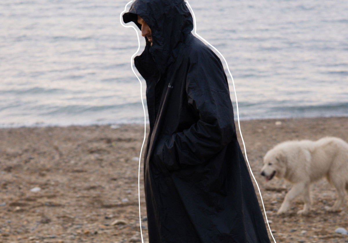 A person wearing a long black VOITED Allweather Coat stands on a pebble beach with the ocean in the background, and a white dog walks nearby. AI modified