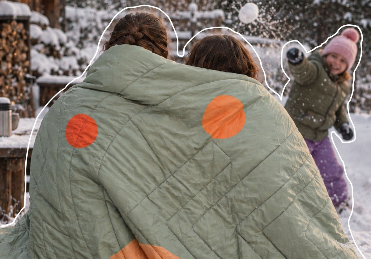 A quilted light sage green Queen Cloudtouch blanket VOITED with two bright orange circular patches, shown from behind in a snowy outdoor setting. Two people are wrapped in it, and a child is playfully throwing snow in the background. AI modified