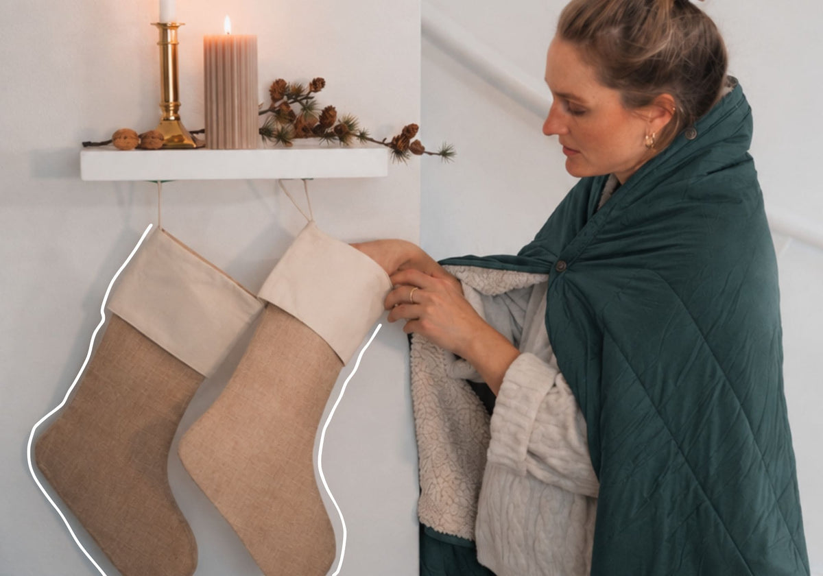 A woman wearing a dark teal green VOITED Cloudtouch blanket is shown in a cozy indoor setting next to two beige burlap Christmas stockings hanging from a white shelf with a candle and pinecones. AI modified