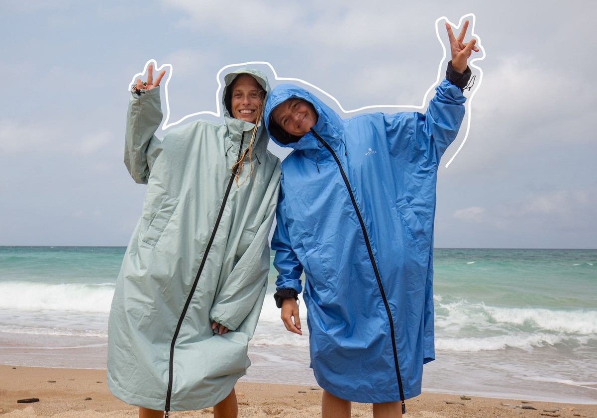 Two people standing on a sandy beach wearing oversized hooded VOITED Allweather Coat, one in light sage green and one in medium blue, with the ocean and cloudy sky behind them.