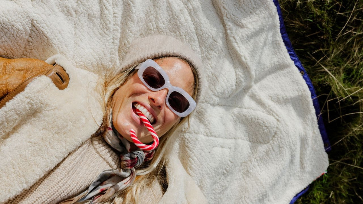 A woman lying on a cream-colored VOITED Cloudtouch blanket outdoors, wearing white cat-eye sunglasses, a beige knit beanie, and a striped knit sweater, with a candy cane in her mouth.