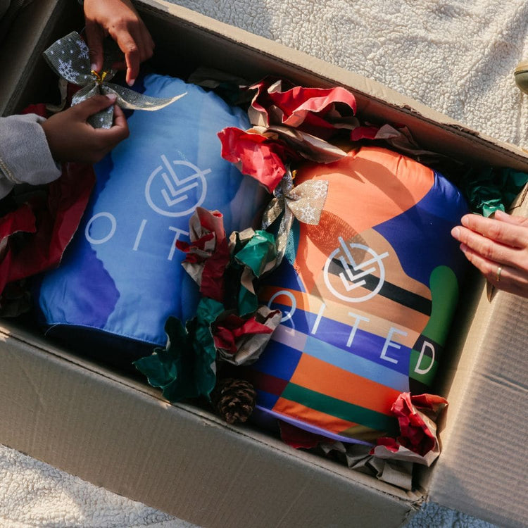 An open cardboard box containing two colorful insulated bags VOITED Cloud touch blankets , surrounded by red and green gift ribbon. Human hands are visible opening the box, indicating a lifestyle or unboxing context.