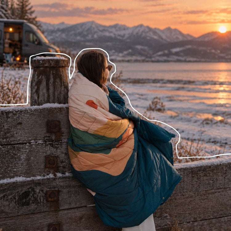 A person sitting outdoors by a snowy lakeside at sunset, wrapped in a VOITED Cloudtouch blanket with horizontal stripes of cream, mustard yellow, teal green, and dark blue. The background shows snow, water, and mountains during golden hour. AI modified