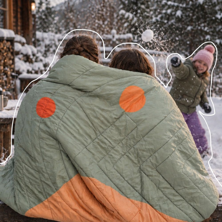 A quilted light sage green Queen Cloudtouch blanket VOITED with two bright orange circular patches, shown from behind in a snowy outdoor setting. Two people are wrapped in it, and a child is playfully throwing snow in the background. AI modified