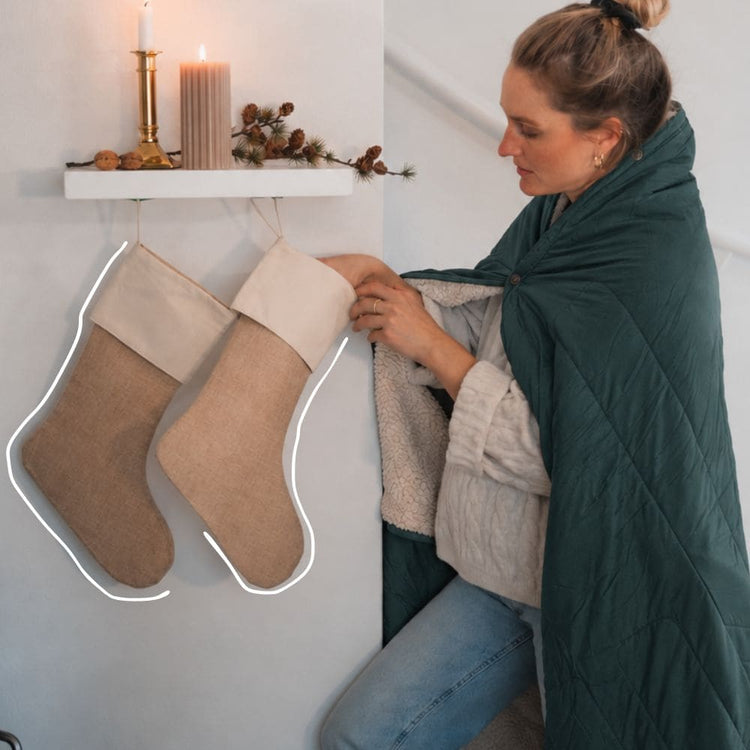 A woman wearing a dark teal green VOITED Cloudtouch blanket is shown in a cozy indoor setting next to two beige burlap Christmas stockings hanging from a white shelf with a candle and pinecones. AI modified