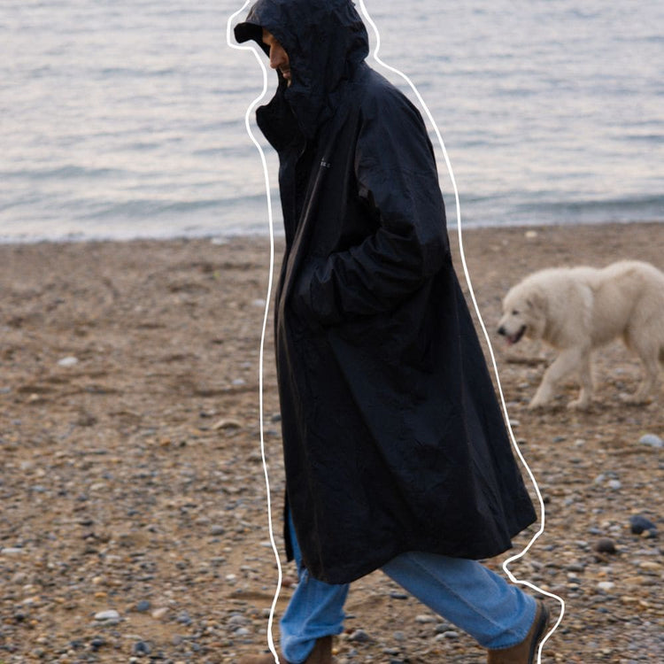 A person wearing a long black VOITED Allweather Coat stands on a pebble beach with the ocean in the background, and a white dog walks nearby. AI modified