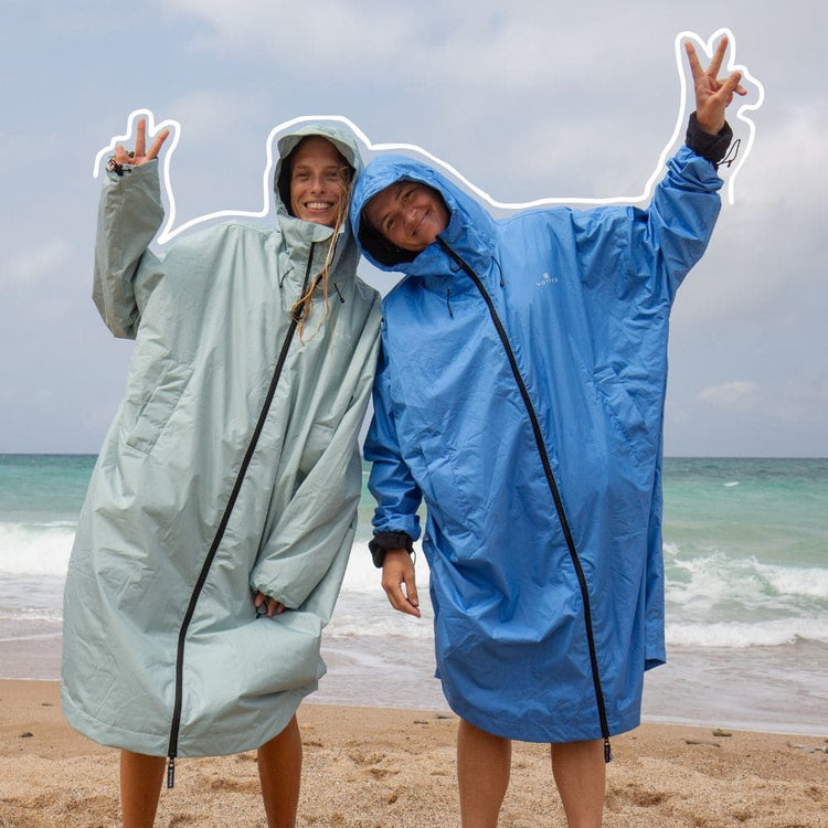 Two people standing on a sandy beach wearing oversized hooded VOITED Allweather Coat, one in light sage green and one in medium blue, with the ocean and cloudy sky behind them.